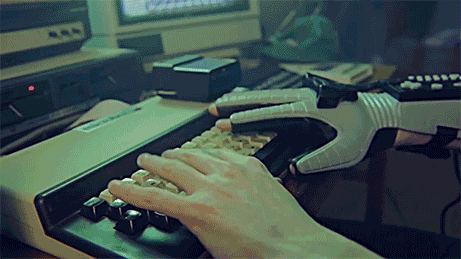 Video stylized to look like a 1980s hacker sequence. Two hands, one of which is wearing a Nintendo Powerglove type randomly at a retro computer console. Cut to the screen, random letters appear rapidly in the middle of some otherwise plausible computer code.