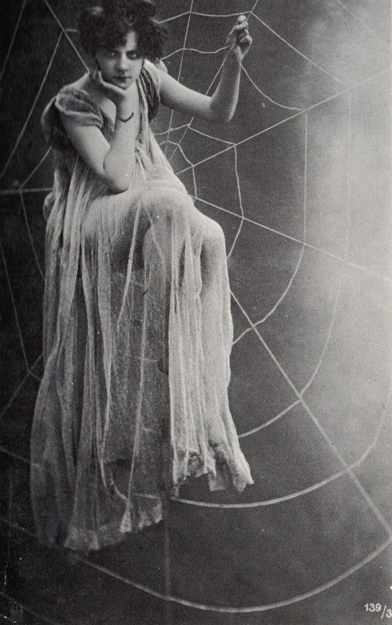 A vintage photo (early 20th century) of a woman in a nightgown sitting suspended in a huge spider web. Her chin is resting on her hand in a slightly pouting gesture.